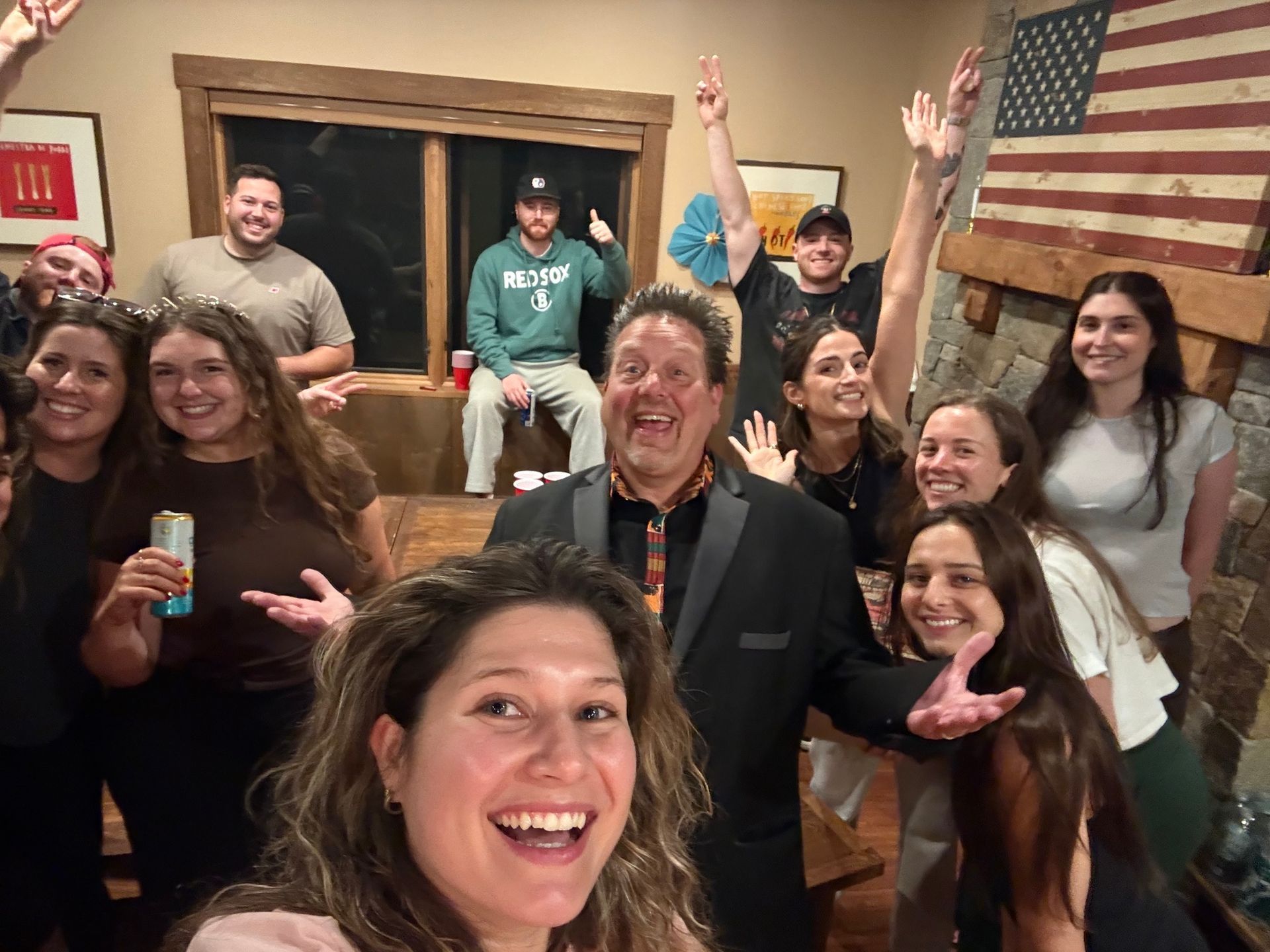 Group of smiling people posing indoors, many waving, with a large American flag on the wall.