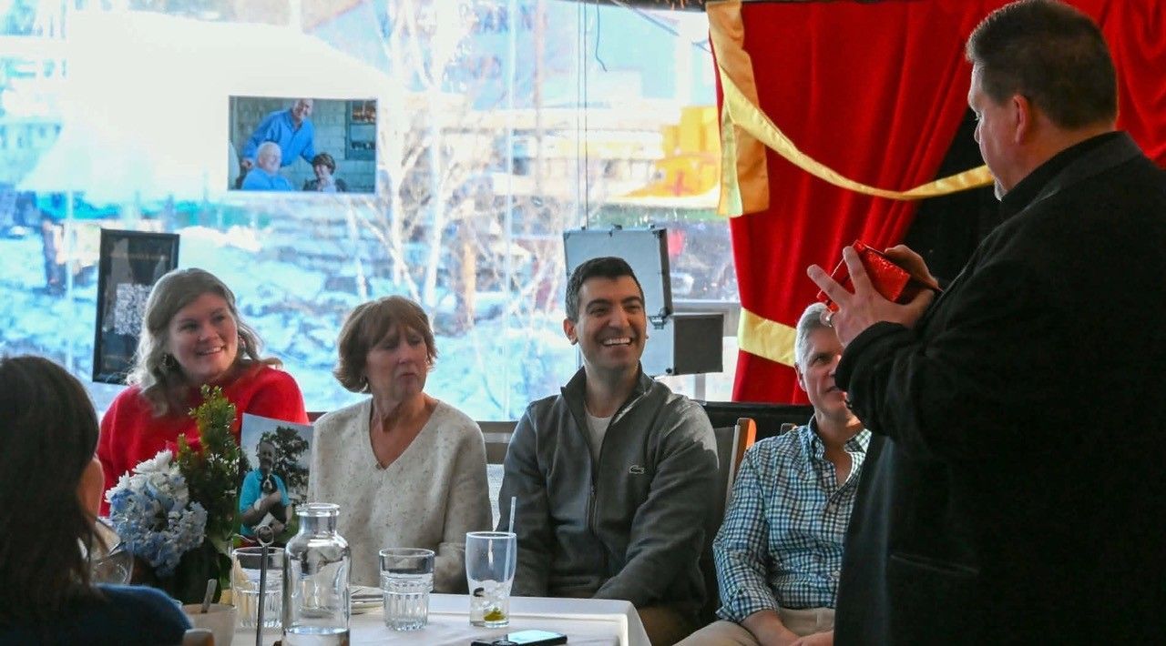 Man speaking to seated guests at a small table in a bright room with red curtains and flowers