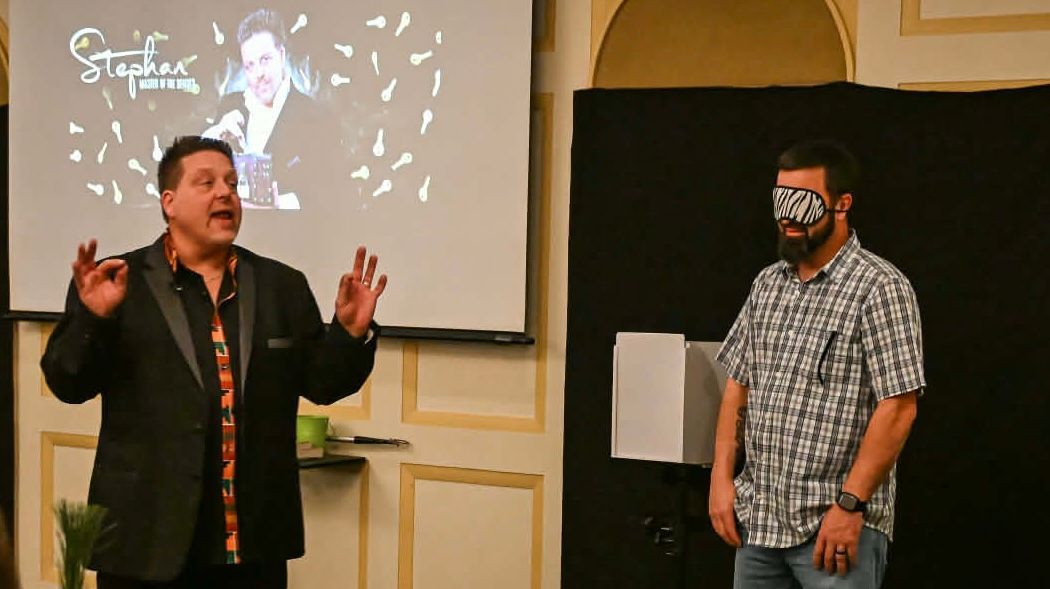 Two men speaking on a stage, one gesturing beside a projected image in the background