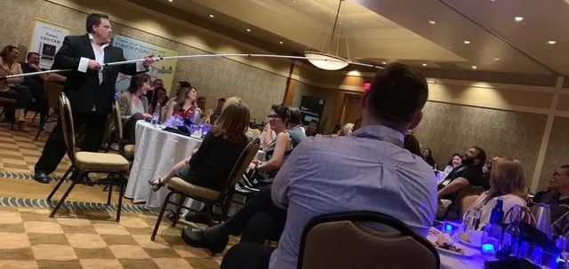 Speaker addressing seated audience at a banquet hall event, with round tables and blue-lit centerpieces.