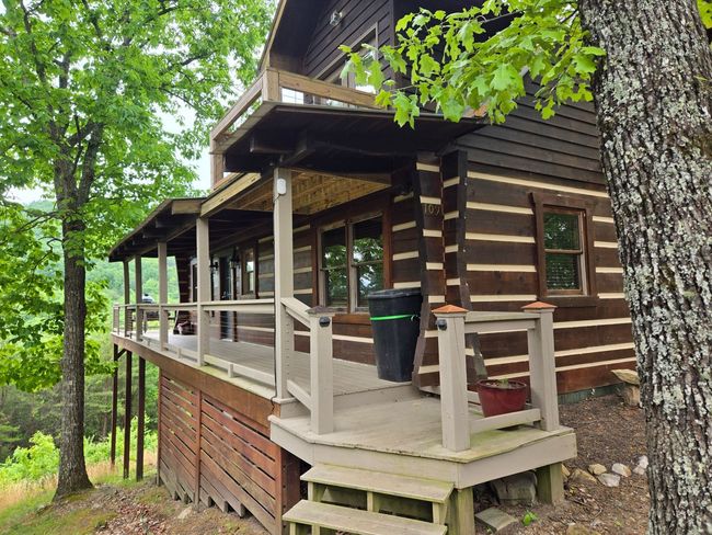 Wooden cabin porch in a wooded setting with railings and steps, surrounded by green trees.