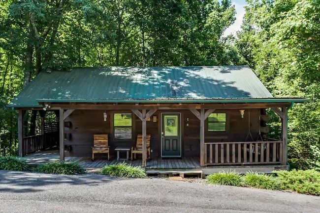 Small rustic log cabin with green roof and front porch, surrounded by trees and gravel drive