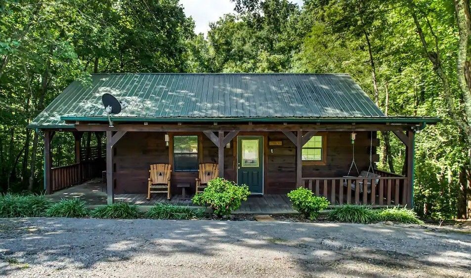 Rustic cabin with green metal roof and wooden porch surrounded by trees and gravel driveway