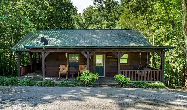 Rustic wooden cabin with green metal roof, porch chairs, and gravel driveway surrounded by trees