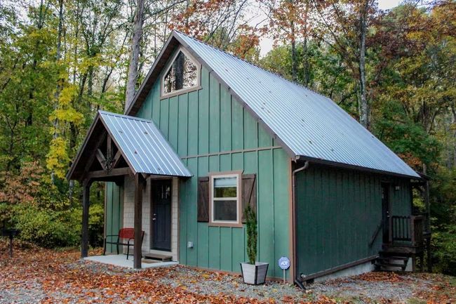 Small green cabin with a blue metal roof and porch, surrounded by autumn trees and fallen leaves.