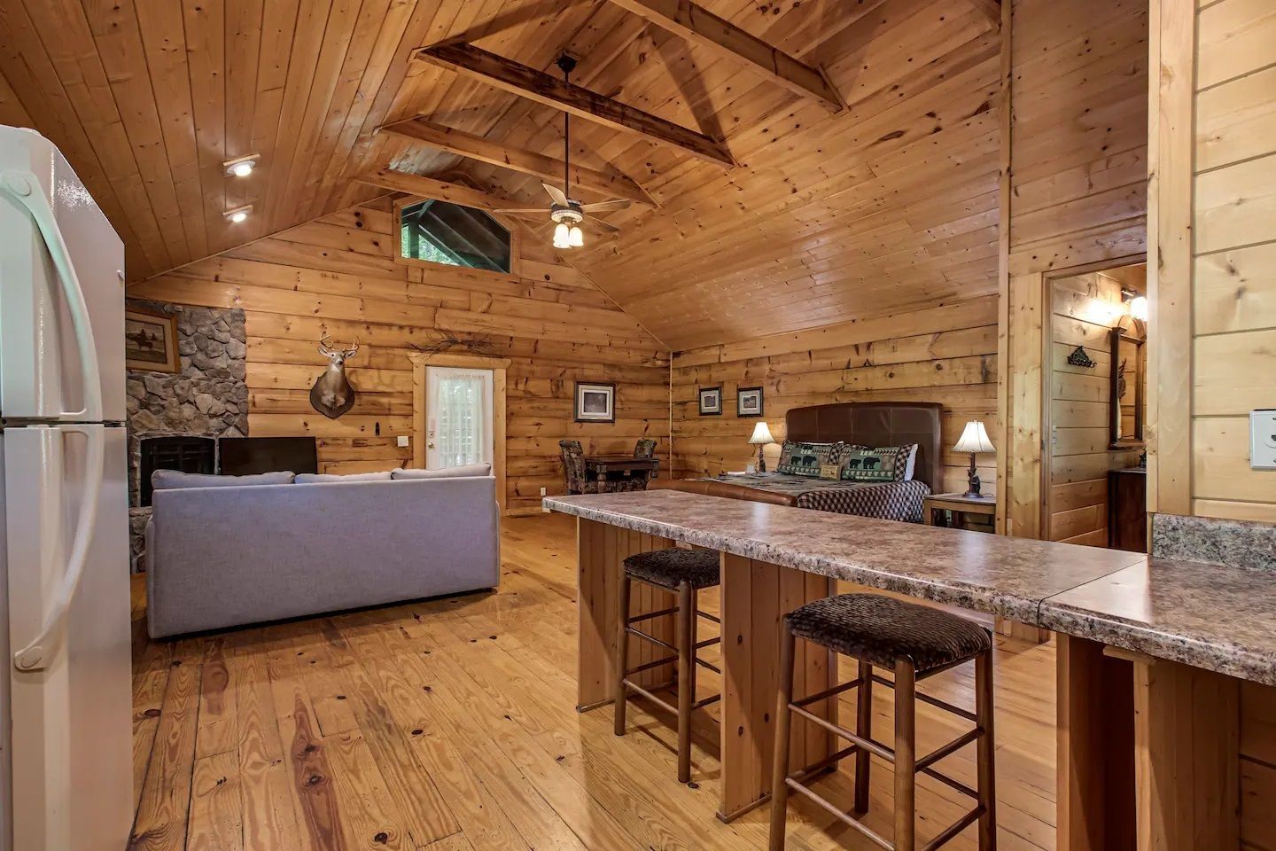 Rustic log cabin kitchen and living area with wood-paneled walls, bar stools, and a granite-topped island.