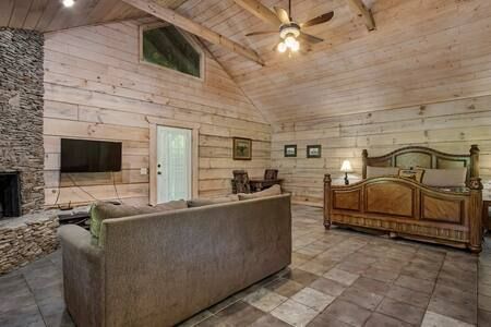 Rustic log cabin bedroom with a king bed, sofa, stone fireplace, and vaulted wood ceiling.