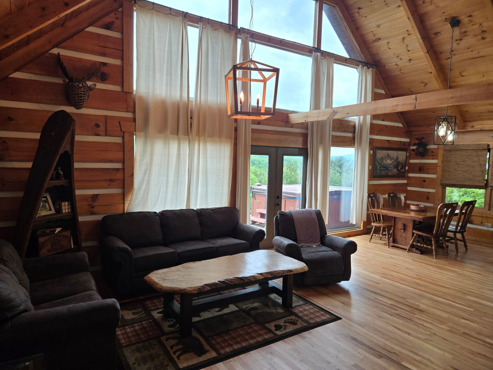 Rustic cabin living room with dark sofas, wood walls, large windows, and dining area beyond
