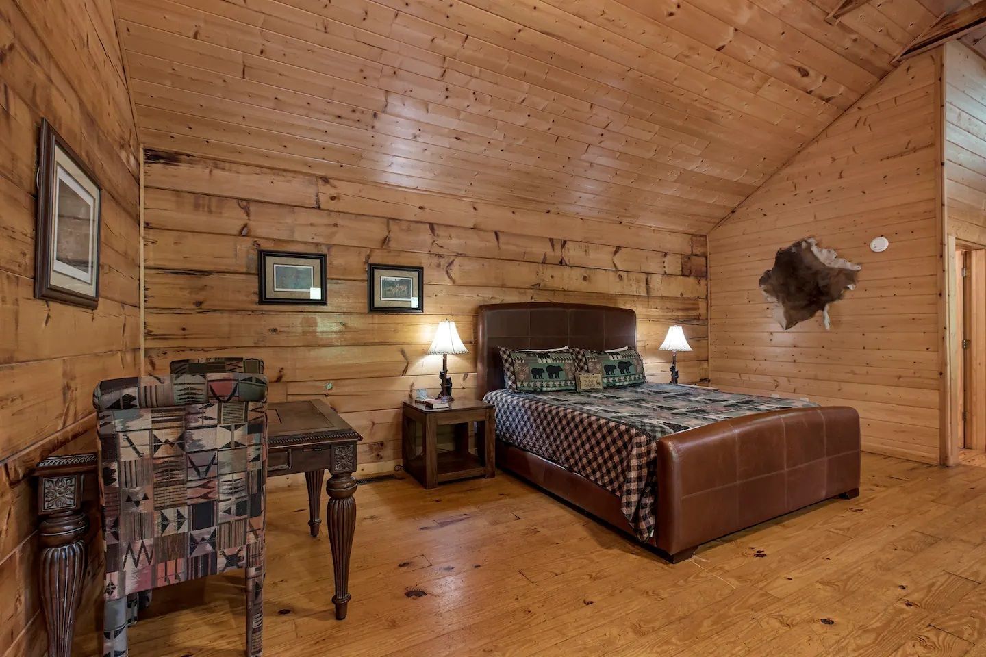 Wood-paneled bedroom with a rustic bed, side table lamp, and patterned chair under sloped wooden ceiling