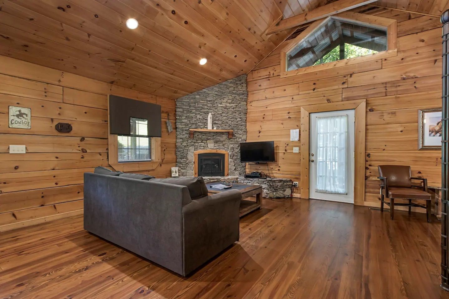 Rustic log cabin living room with stone fireplace, dark sofa, wood floors, and vaulted ceiling