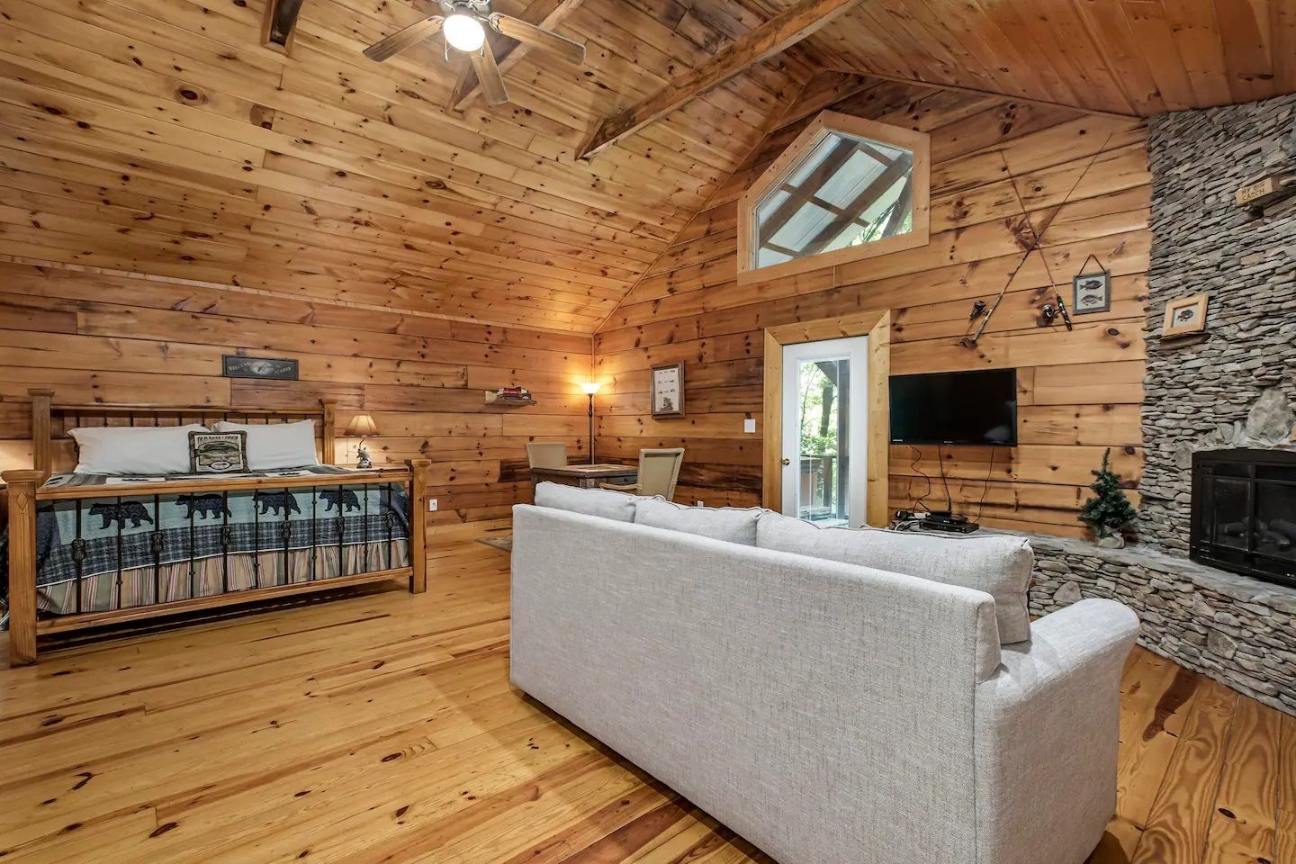 Rustic log cabin living room with stone fireplace, TV, and light gray sofa under vaulted wood ceiling