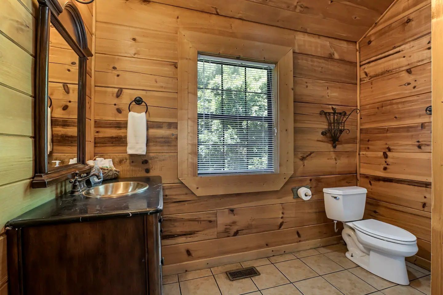 Rustic wood-paneled bathroom with a sink, mirror, window, and toilet on tile floor