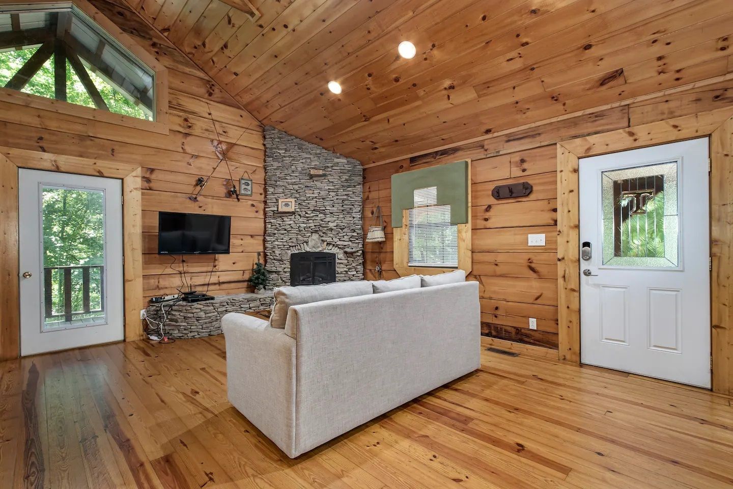 Cozy wood-paneled cabin living room with stone fireplace, white sofa, and windows on both sides