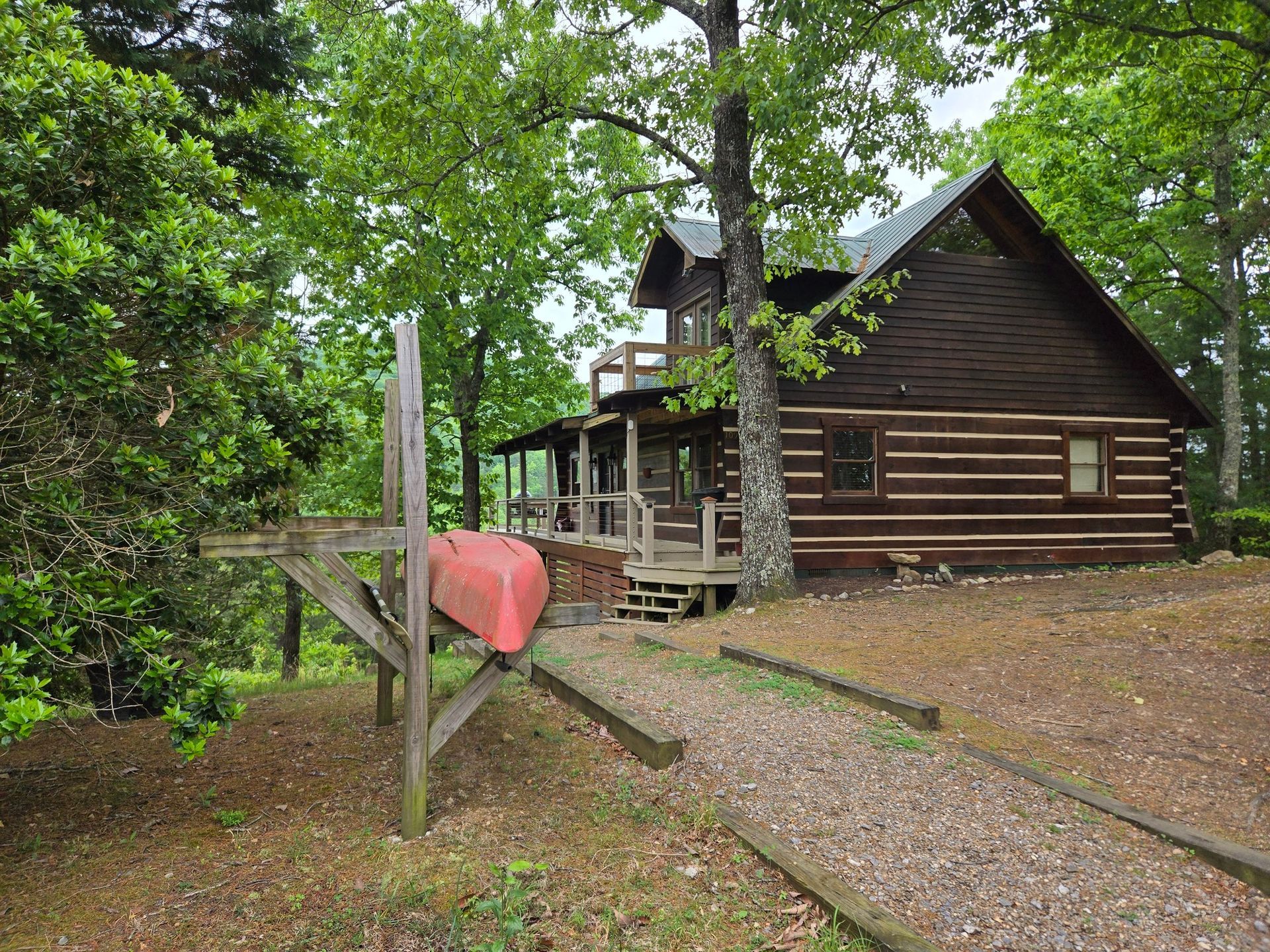 Rustic log cabin in a wooded area with a wooden footbridge and a red heart-shaped sign in front