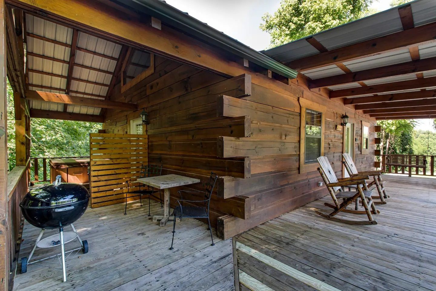 Covered wooden patio with grill, table, and folding chairs beside a rustic cabin and deck