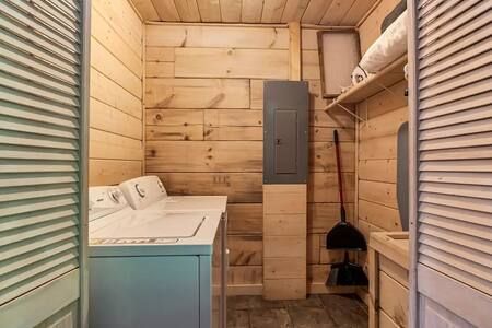 Small laundry room with a washer, shelf, broom, and louvered doors in a wooden interior.