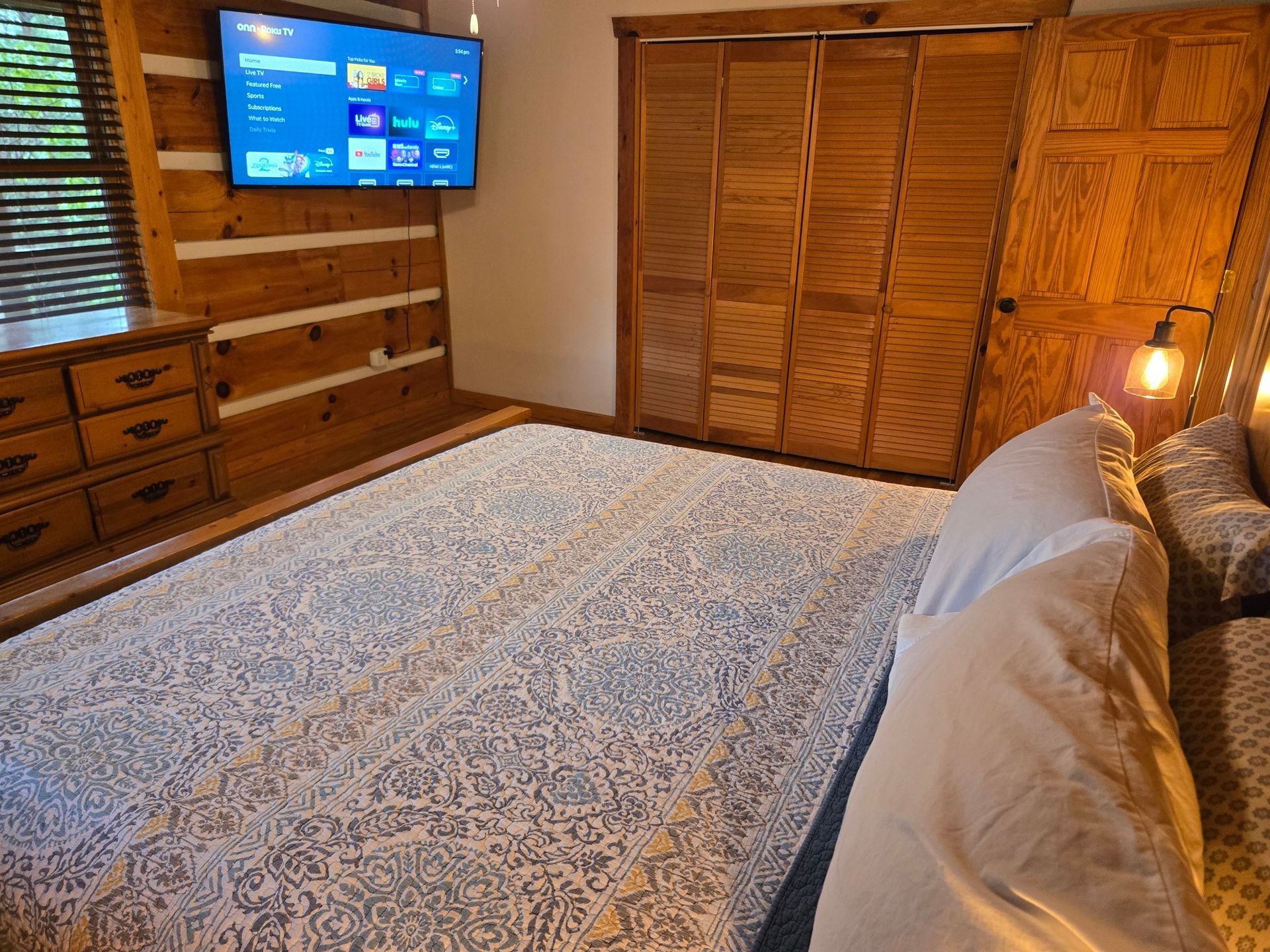 Bedroom with a wall-mounted TV, wooden dresser, closet doors, and a neatly made bed with patterned bedding.
