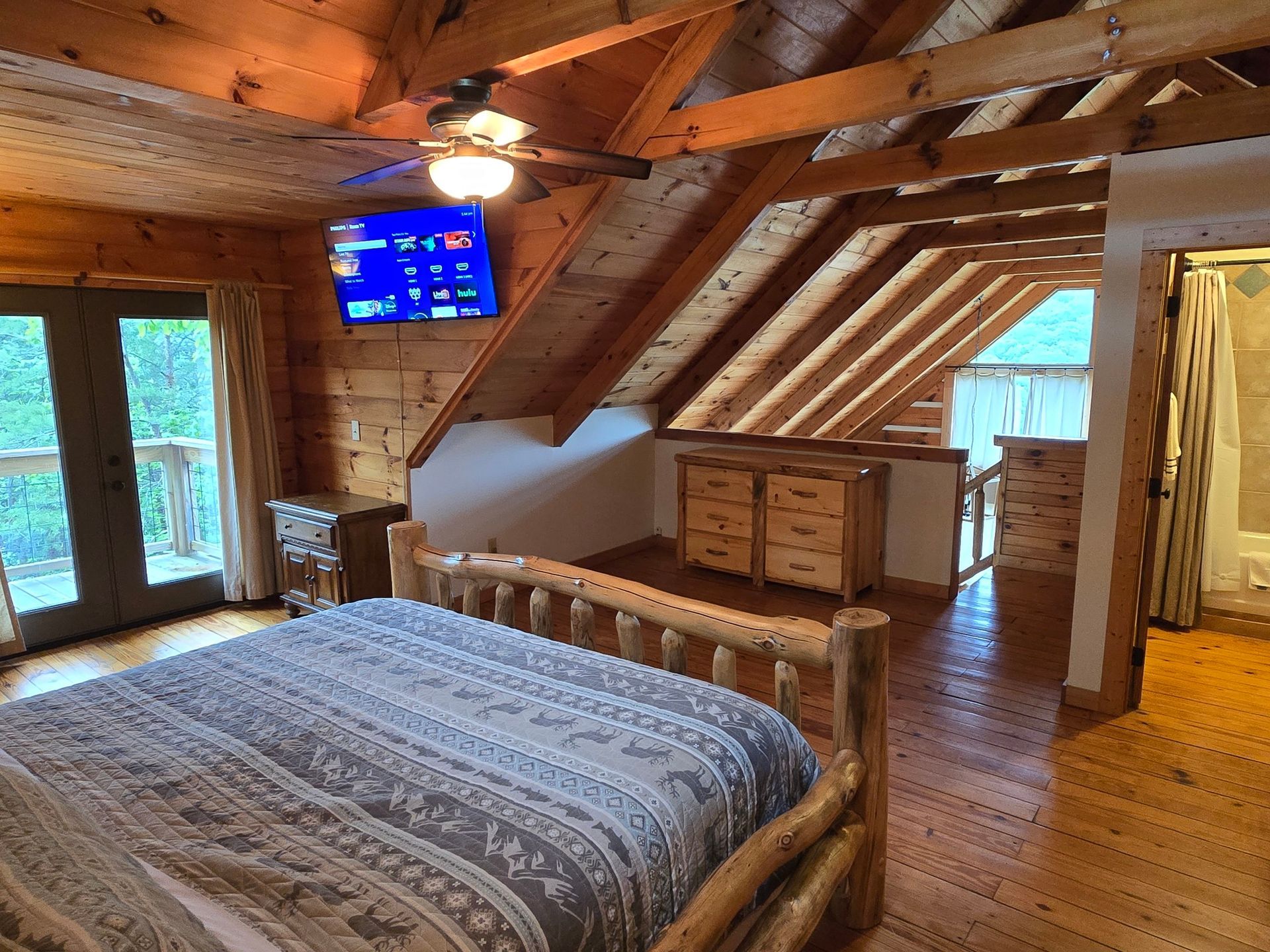 Cozy attic bedroom with a wooden bed, sloped ceiling, TV, dresser, and windows overlooking trees