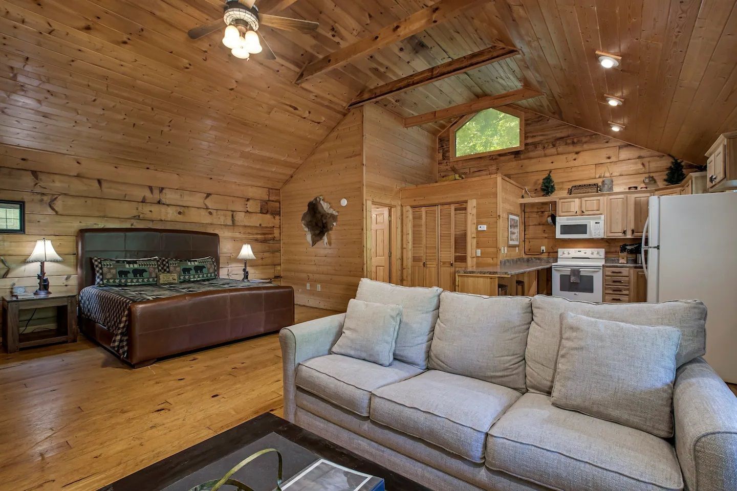 Cozy log cabin bedroom with vaulted wood ceiling, queen bed, sofa, and kitchenette area