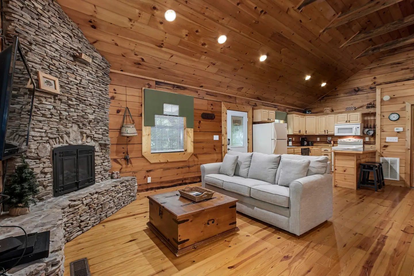 Cozy rustic living room with stone fireplace, wood walls and ceiling, and a gray sofa on hardwood floors