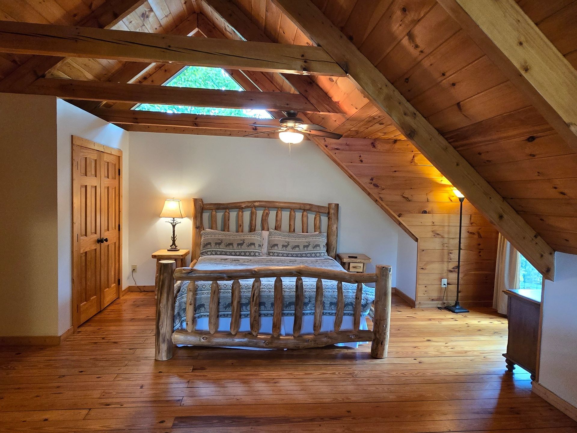 Cozy attic bedroom with wooden beams, a bed, nightstand, and slanted ceiling windows.