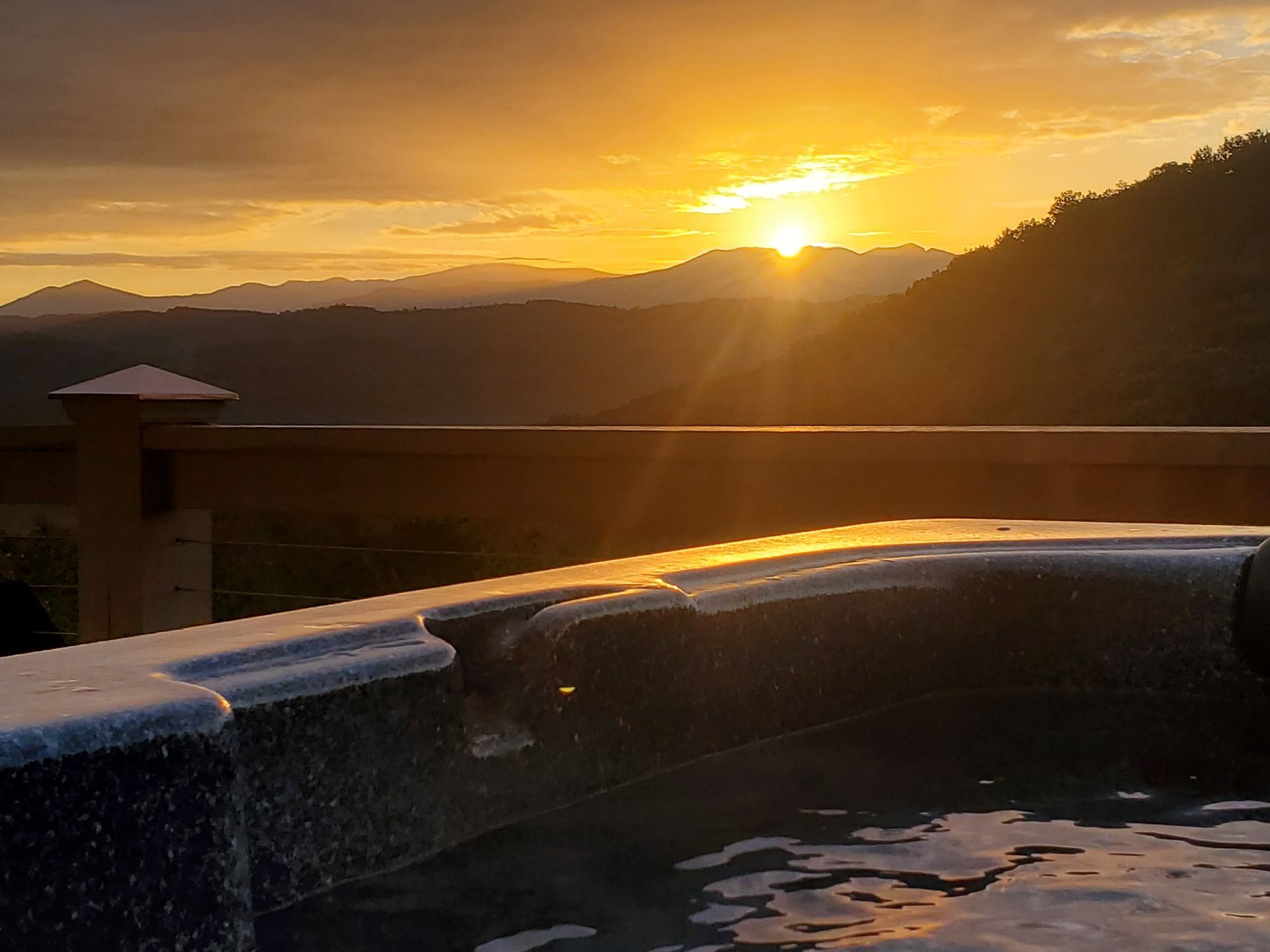 Sunset over mountains viewed from a hot tub with rippling water and a deck railing