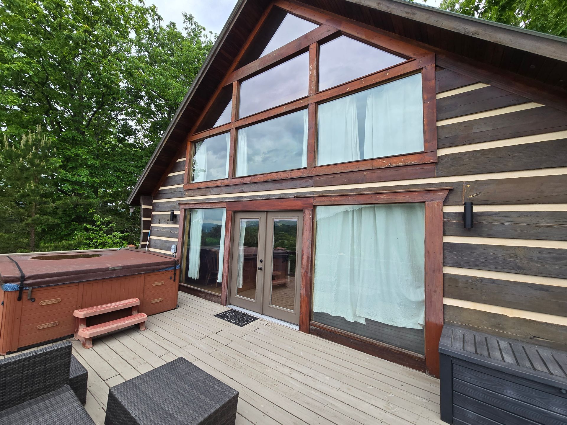 Wooden cabin deck with hot tub and lounge chairs beneath large windows and trees