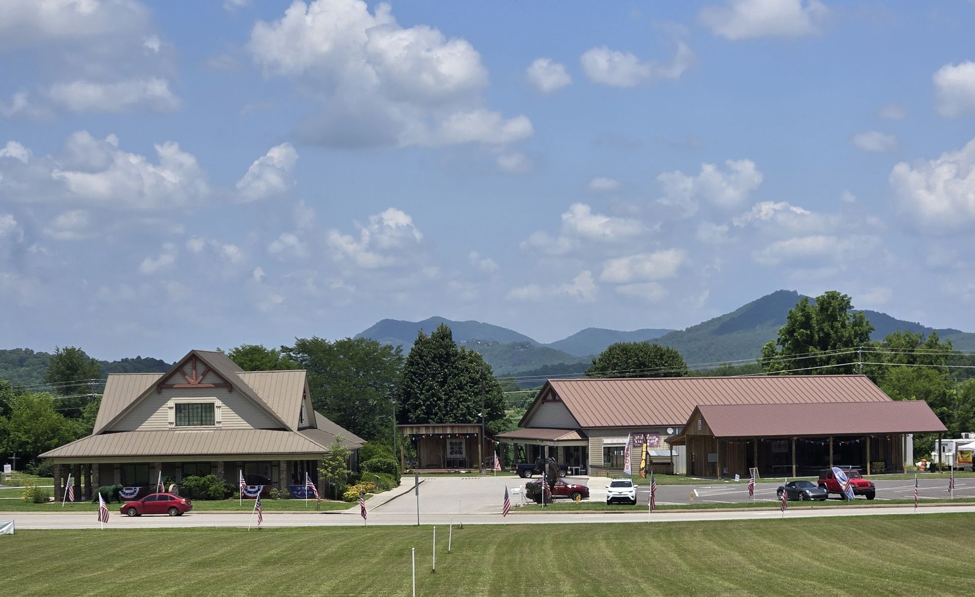 Two buildings with parked cars in front, set against green mountains and a partly cloudy sky.