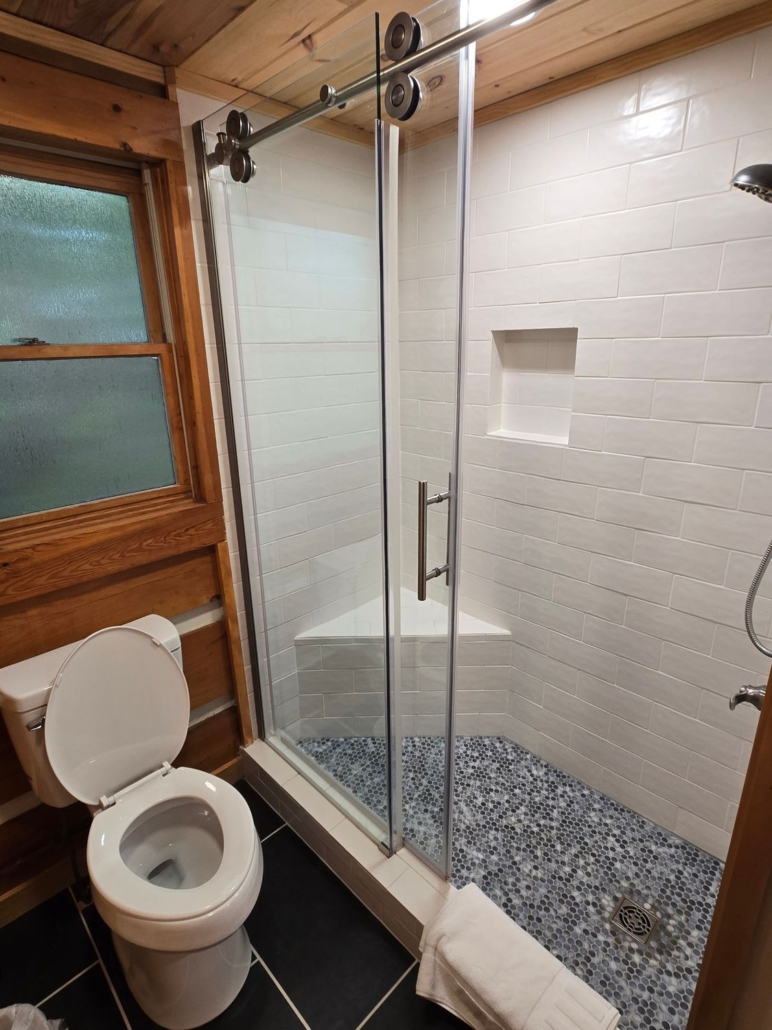 Compact bathroom with toilet and glass shower enclosure, white tiled walls, black floor, and wooden ceiling.