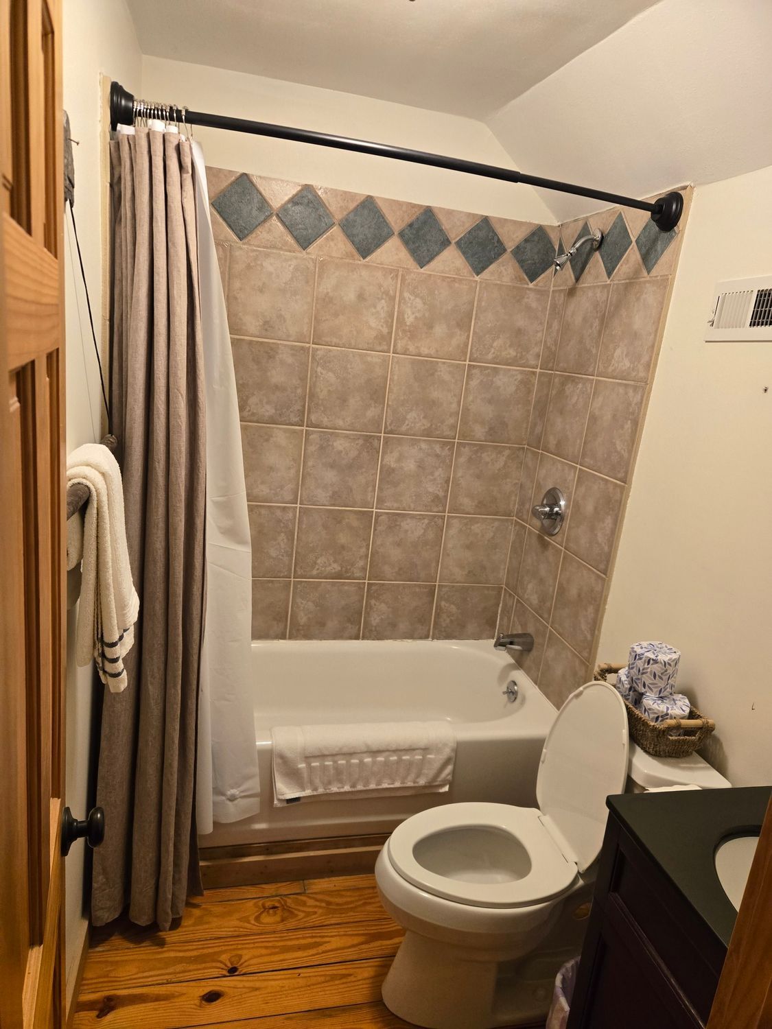 Small bathroom with tub and shower curtain, beige tiled walls, wooden floor, and a toilet in the foreground