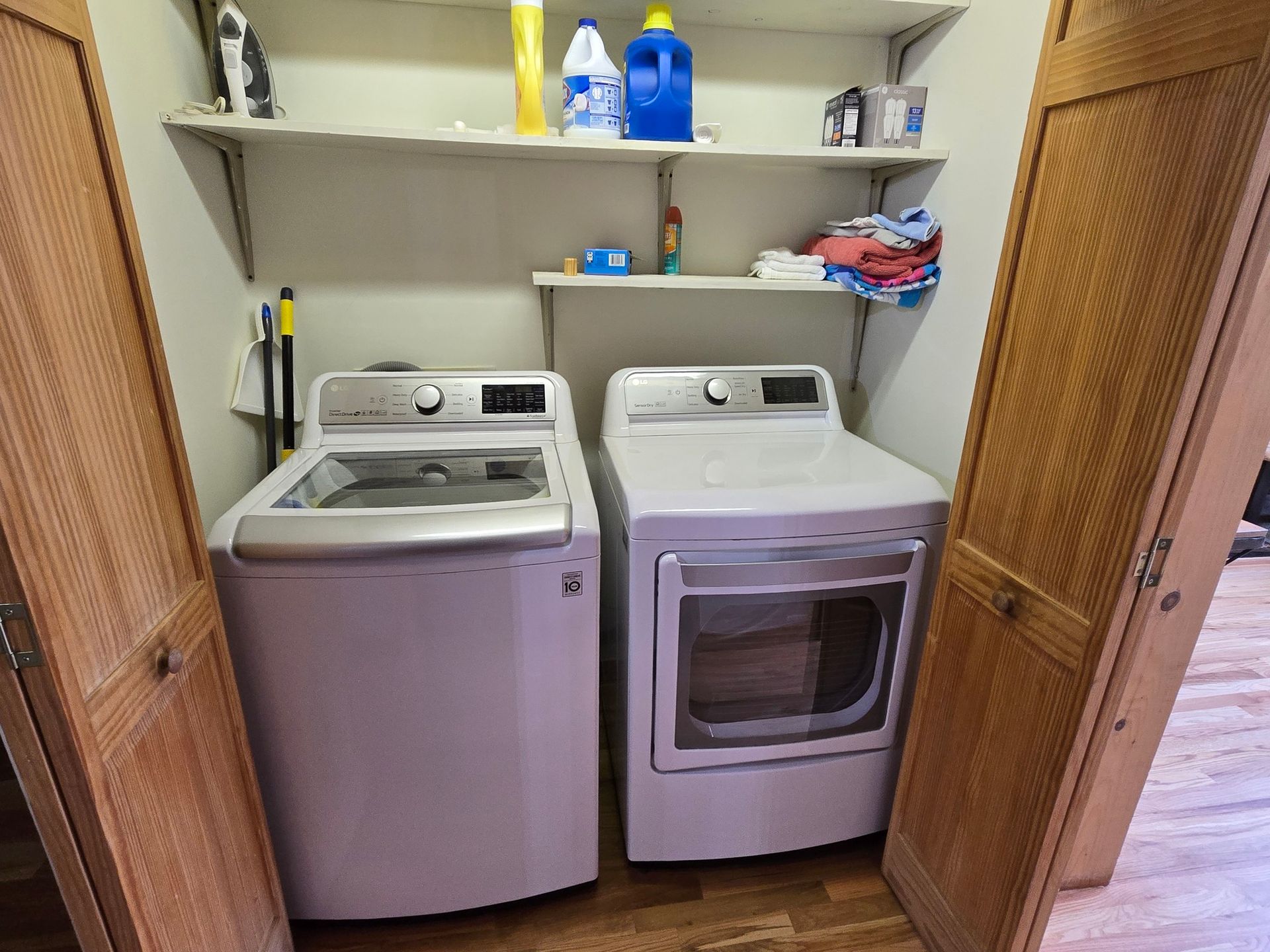 Laundry closet with a white top-load washer and matching front-load dryer beneath shelves of cleaning supplies