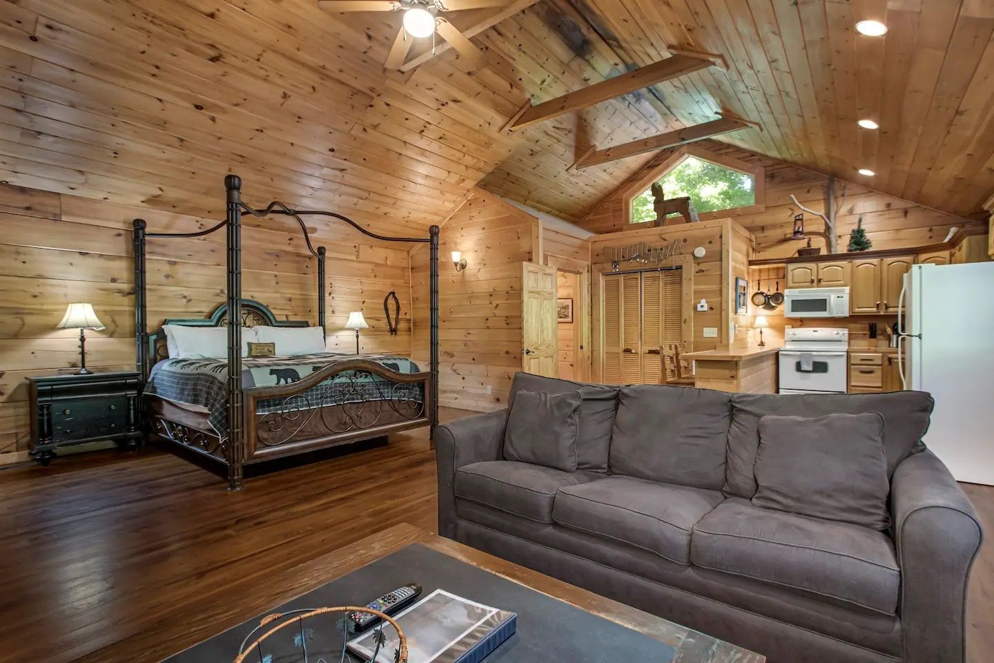 Cozy rustic loft bedroom with wood-paneled ceiling, black four-poster bed, gray sofa, and kitchen area