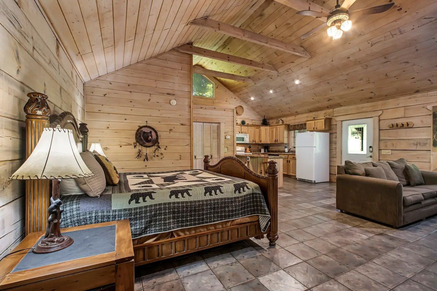 Cozy log cabin bedroom with a wooden bed, lamp, sofa, tile floor, and kitchenette area