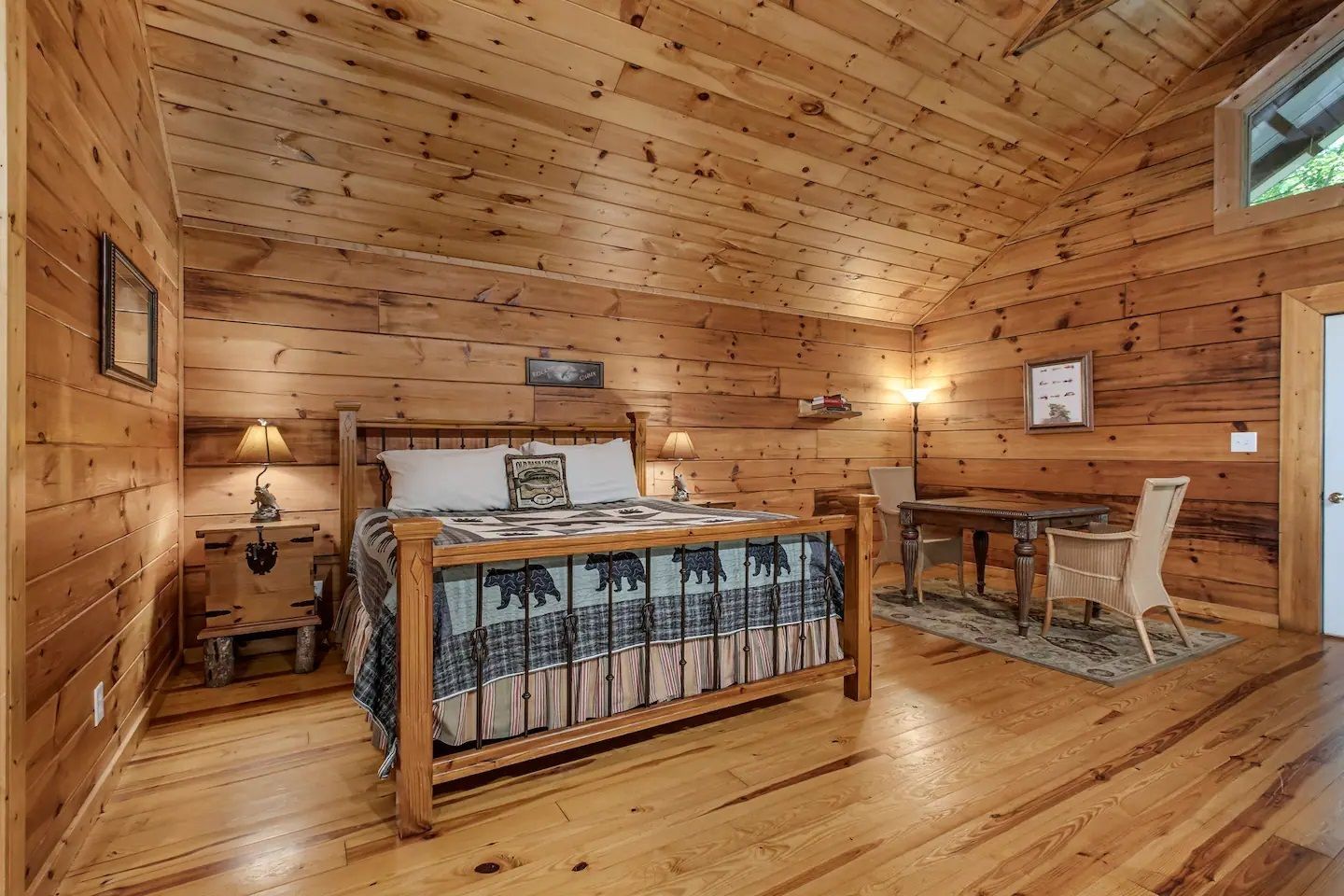 Cozy wooden bedroom with a bed, lamp, desk, chair, and warm rustic cabin interior