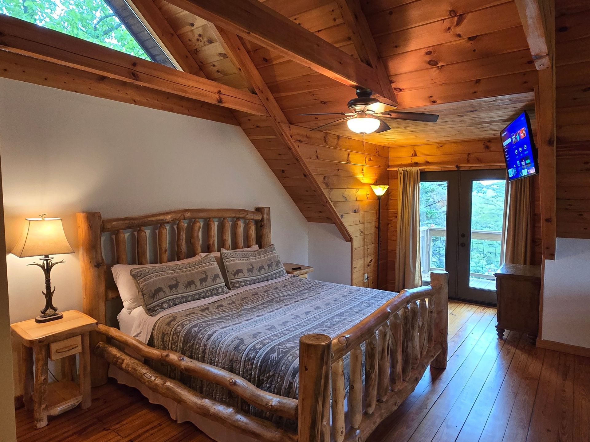Cozy log cabin bedroom with wooden bed, sloped ceiling, lamp, and large window overlooking trees