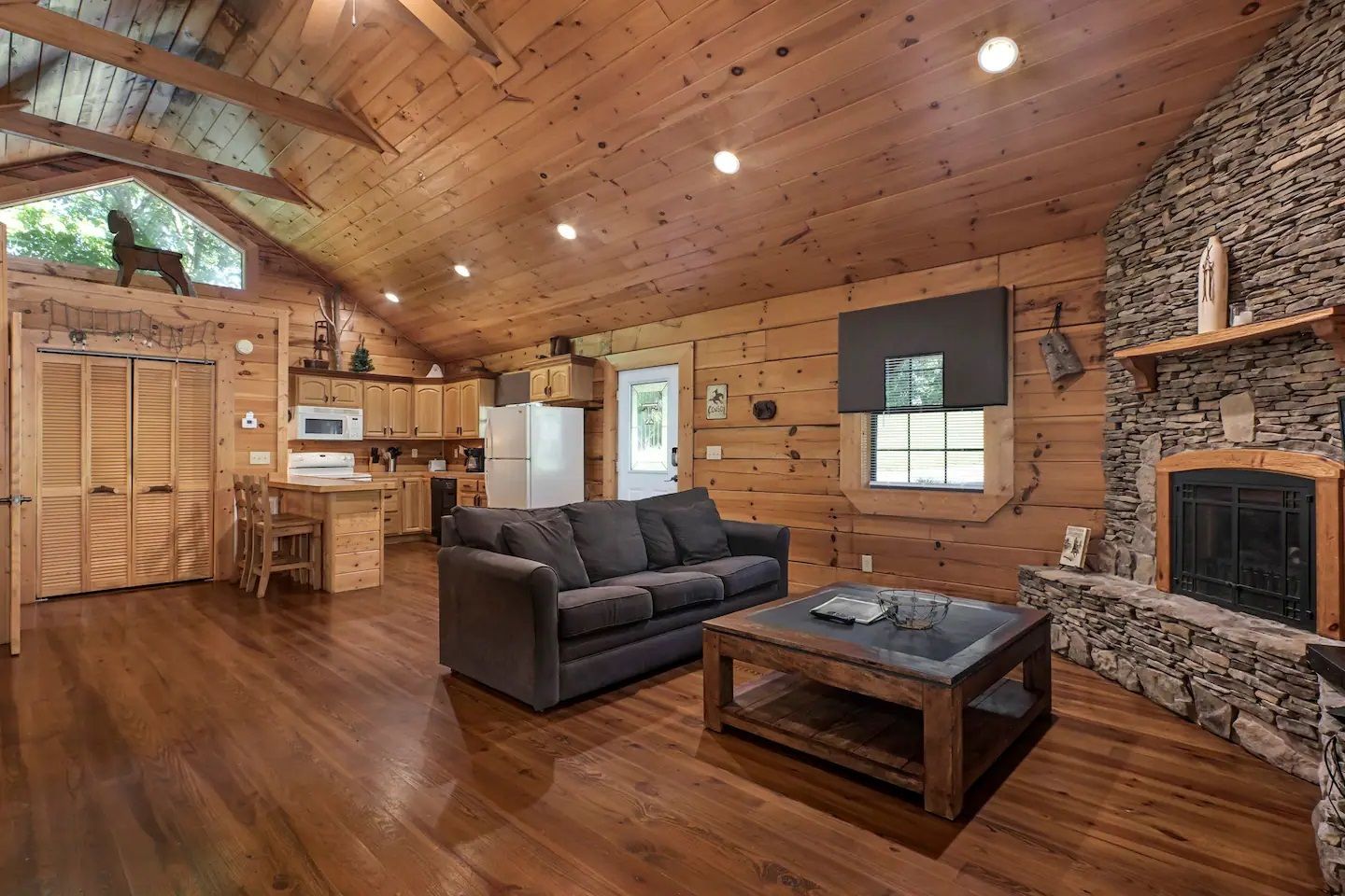 Rustic wood-paneled living room with a stone fireplace, gray sofa, and kitchen area in the background