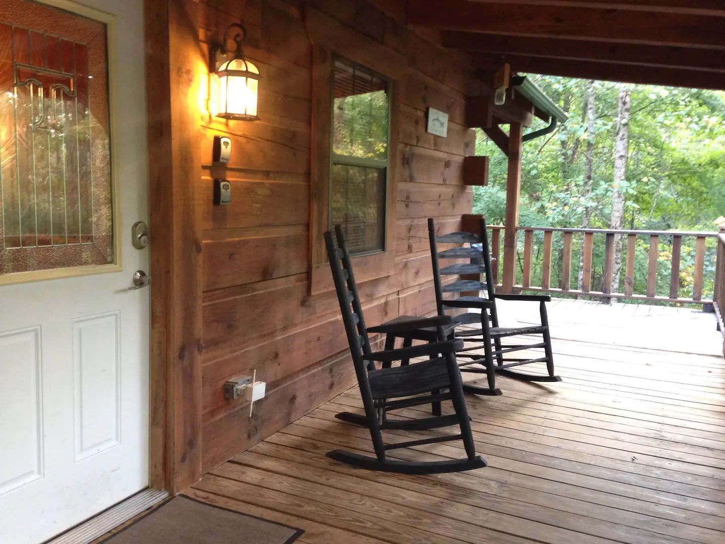Wooden porch with two rocking chairs, warm wall lantern, and white door beside a forest view.