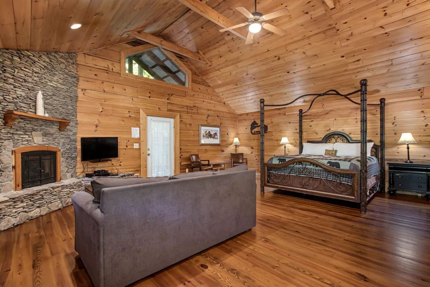 Cozy log cabin living room with stone fireplace, gray sofa, and four-poster bed, wood-paneled walls and ceiling
