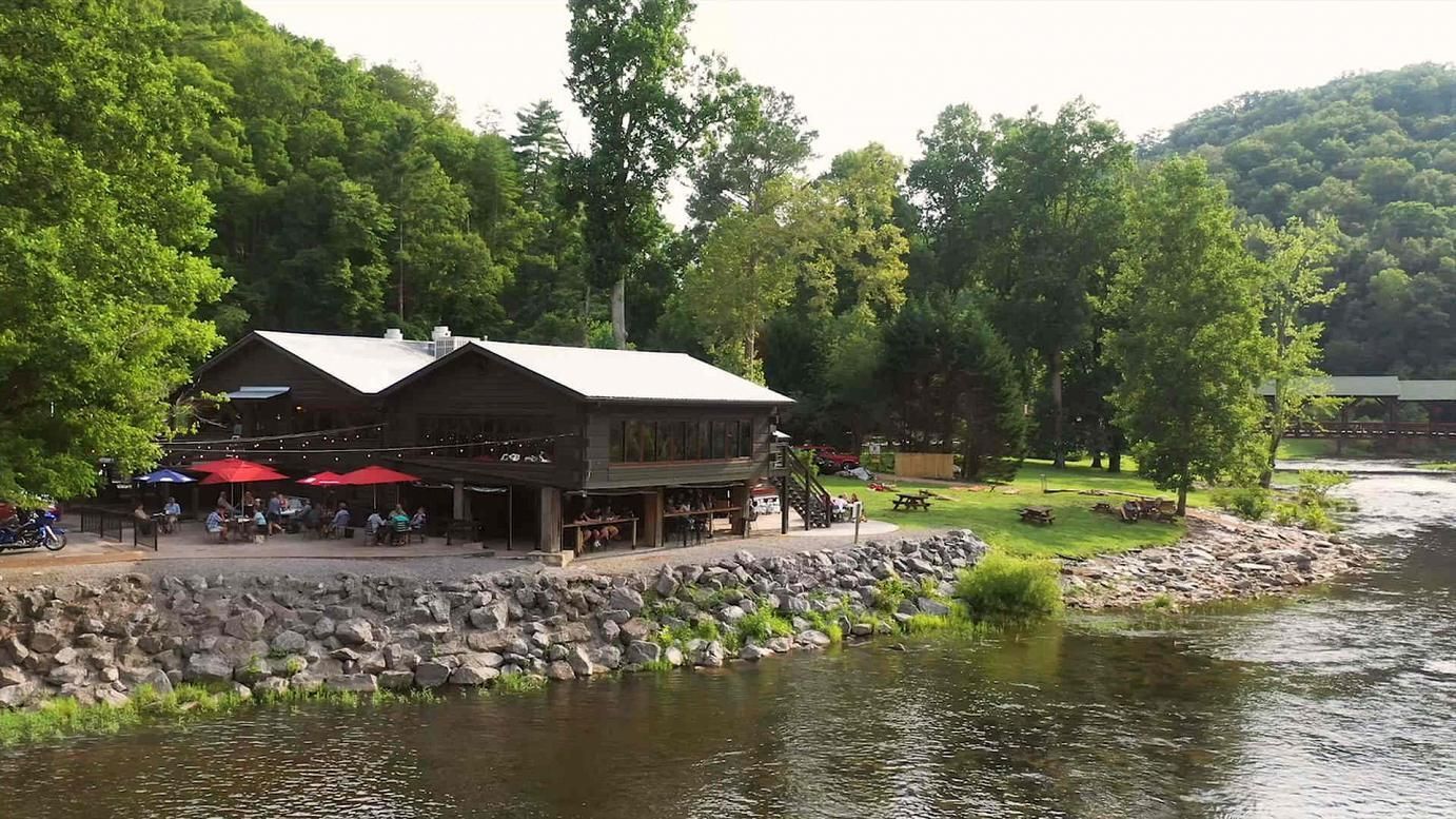 Lakeside restaurant with a red umbrella terrace beside a calm river, surrounded by lush green trees