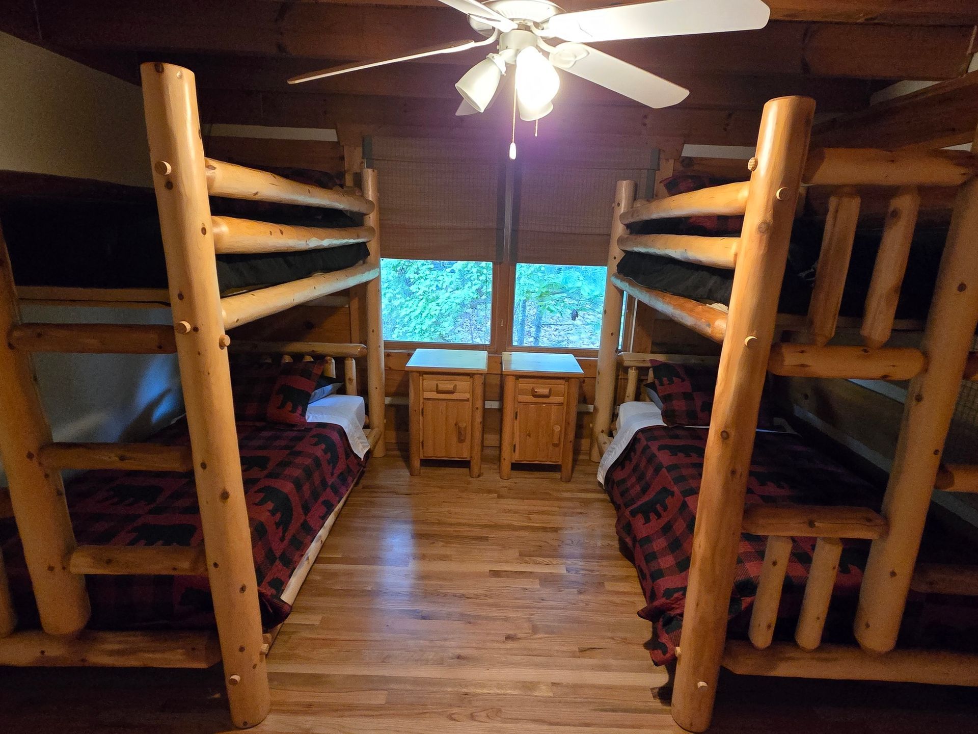 Wooden cabin bedroom with two bunk beds, a ceiling fan, and a window above twin bedside cabinets.
