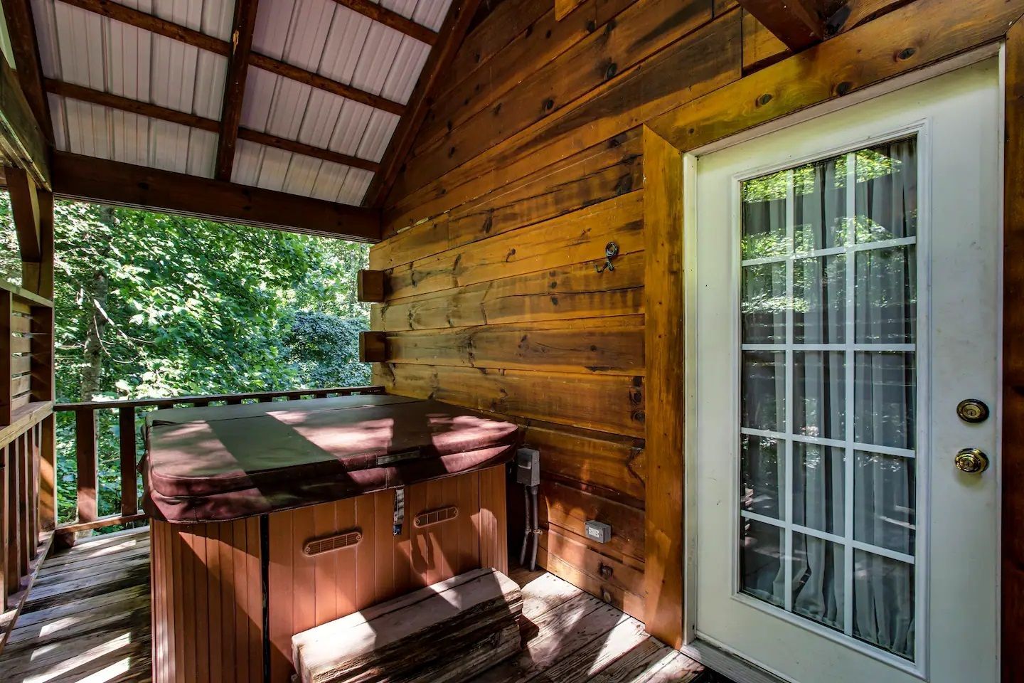 Covered wooden porch with a hot tub beside a glass door and railing overlooking trees