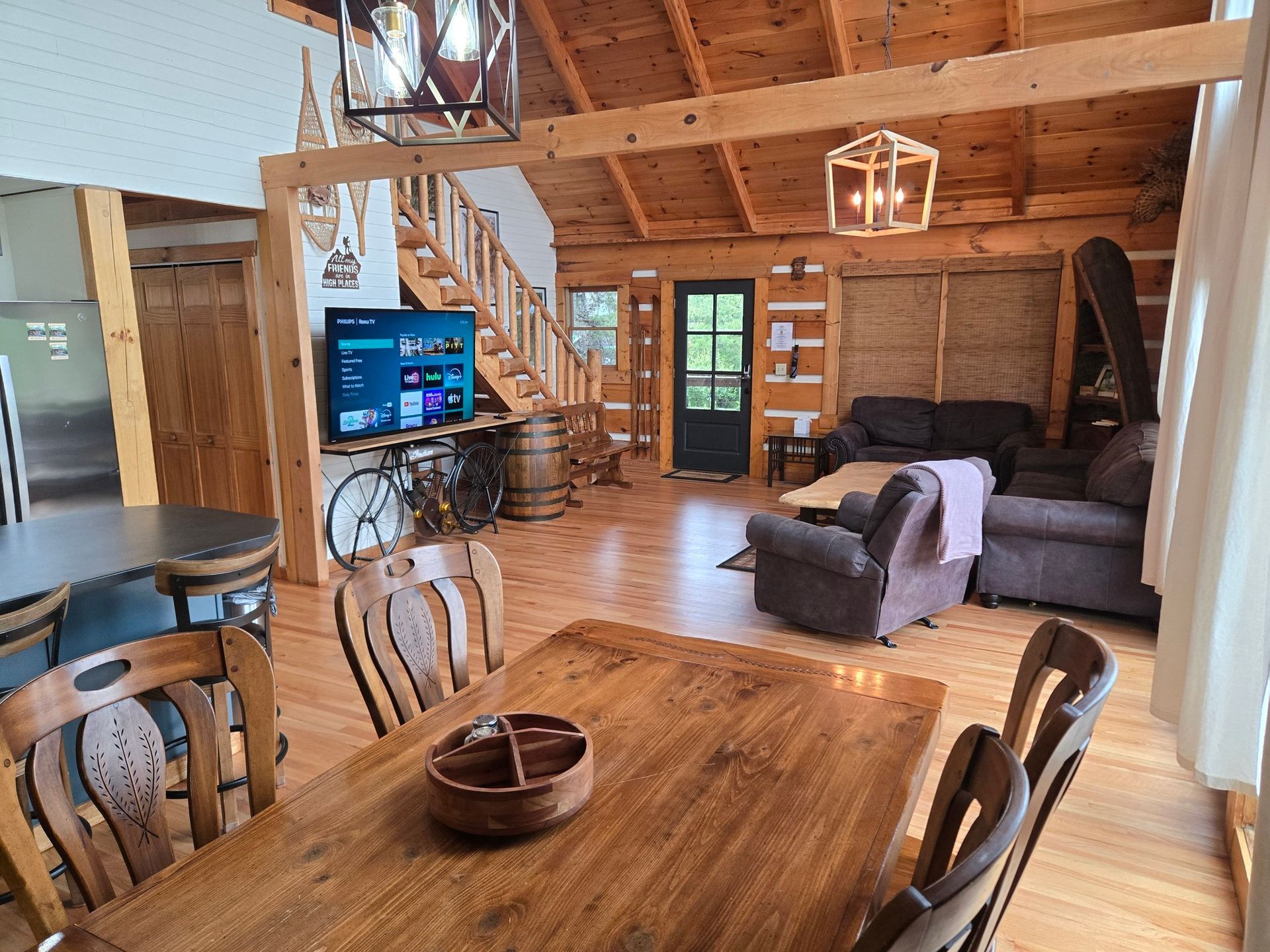Rustic cabin living room with wood beams, dining table, stairs, TV, and two armchairs.