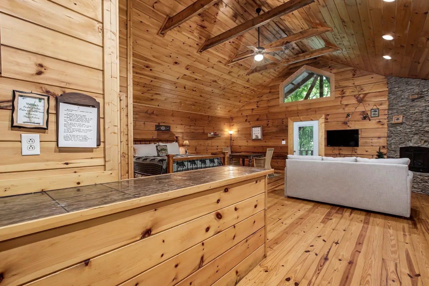 Rustic wood-paneled cabin interior with vaulted ceiling, kitchen area, and gray sofa under warm lighting
