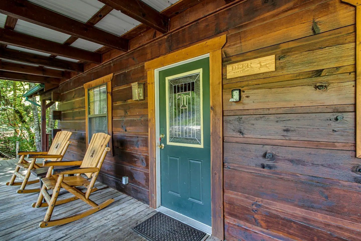 Wooden cabin porch with two chairs, a green door, and a woven rug under a covered roof.