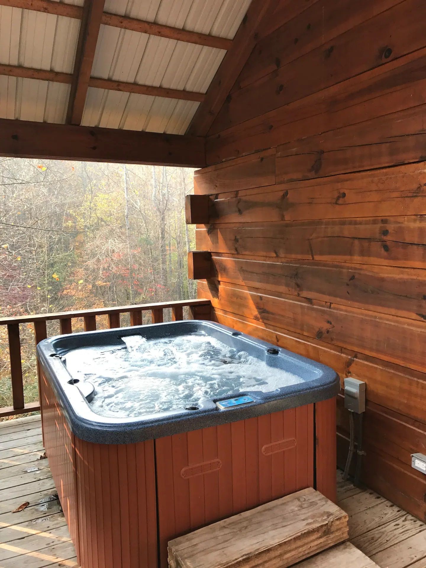 Hot tub on a wooden deck under a covered porch, with a wooded view beyond the railing.