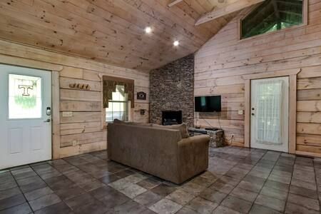 Cozy rustic living room with wood-paneled walls, stone fireplace, tiled floor, and a brown sofa