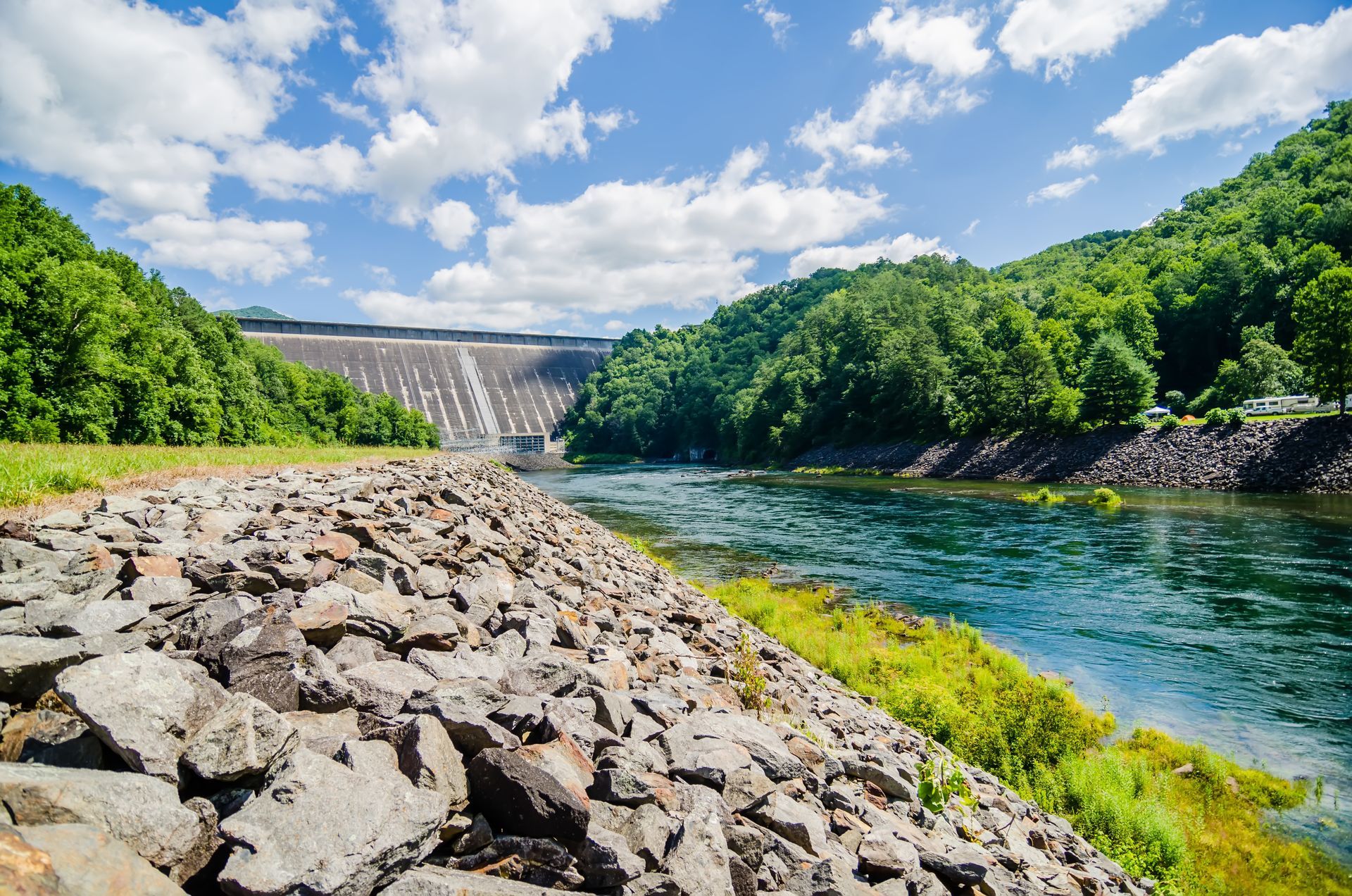 Rocky riverbank beside a blue river, with a dam and green forest under a bright cloudy sky