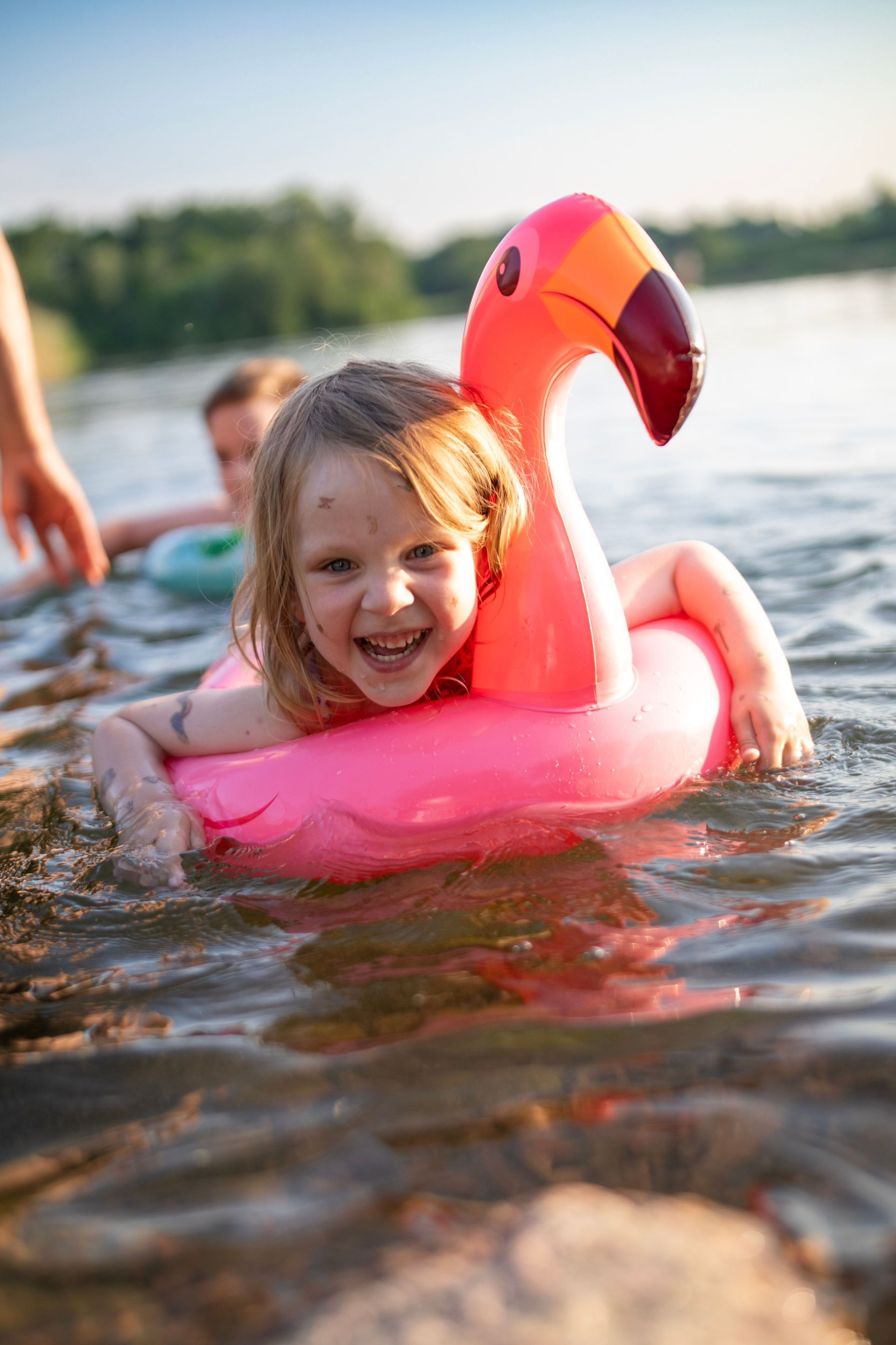 Child smiling on a pink flamingo float in a lake, with splashing water and a blurred shoreline behind.