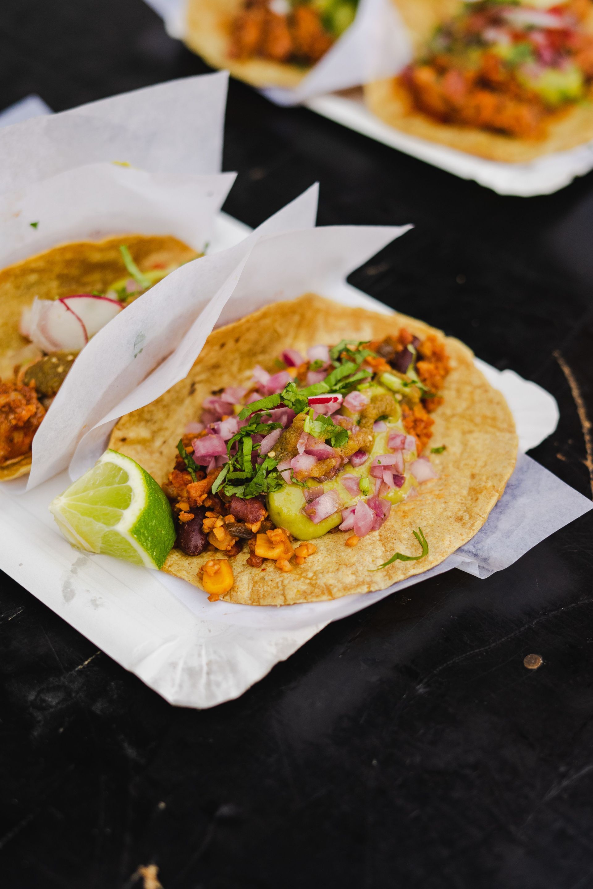 Two tacos on white parchment with lime wedges, topped with onions and herbs on a dark table