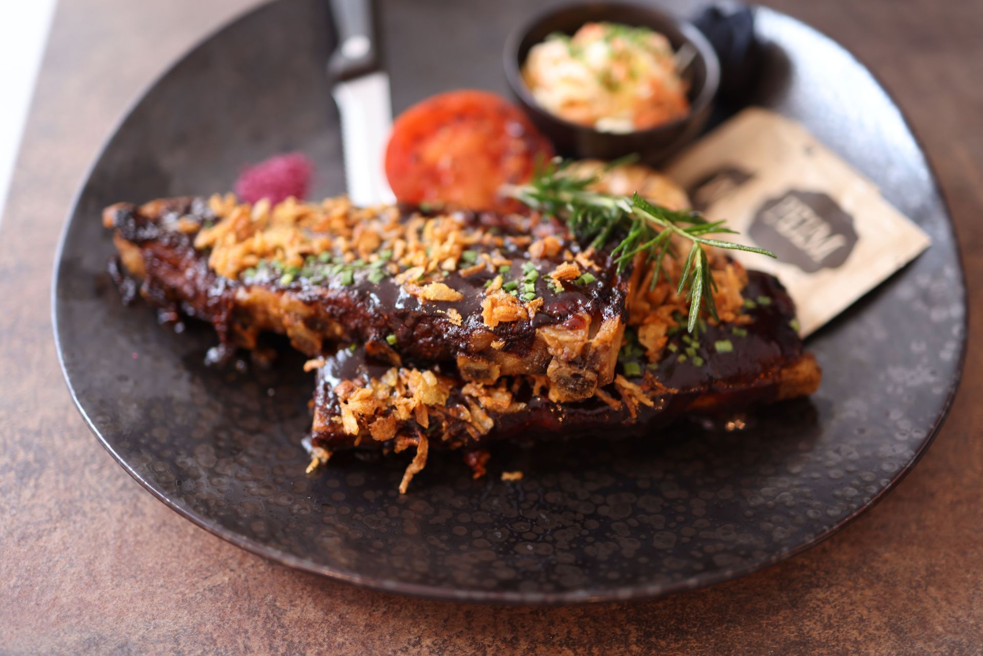 Charred glazed ribs on a black plate with garnish and two small side dishes